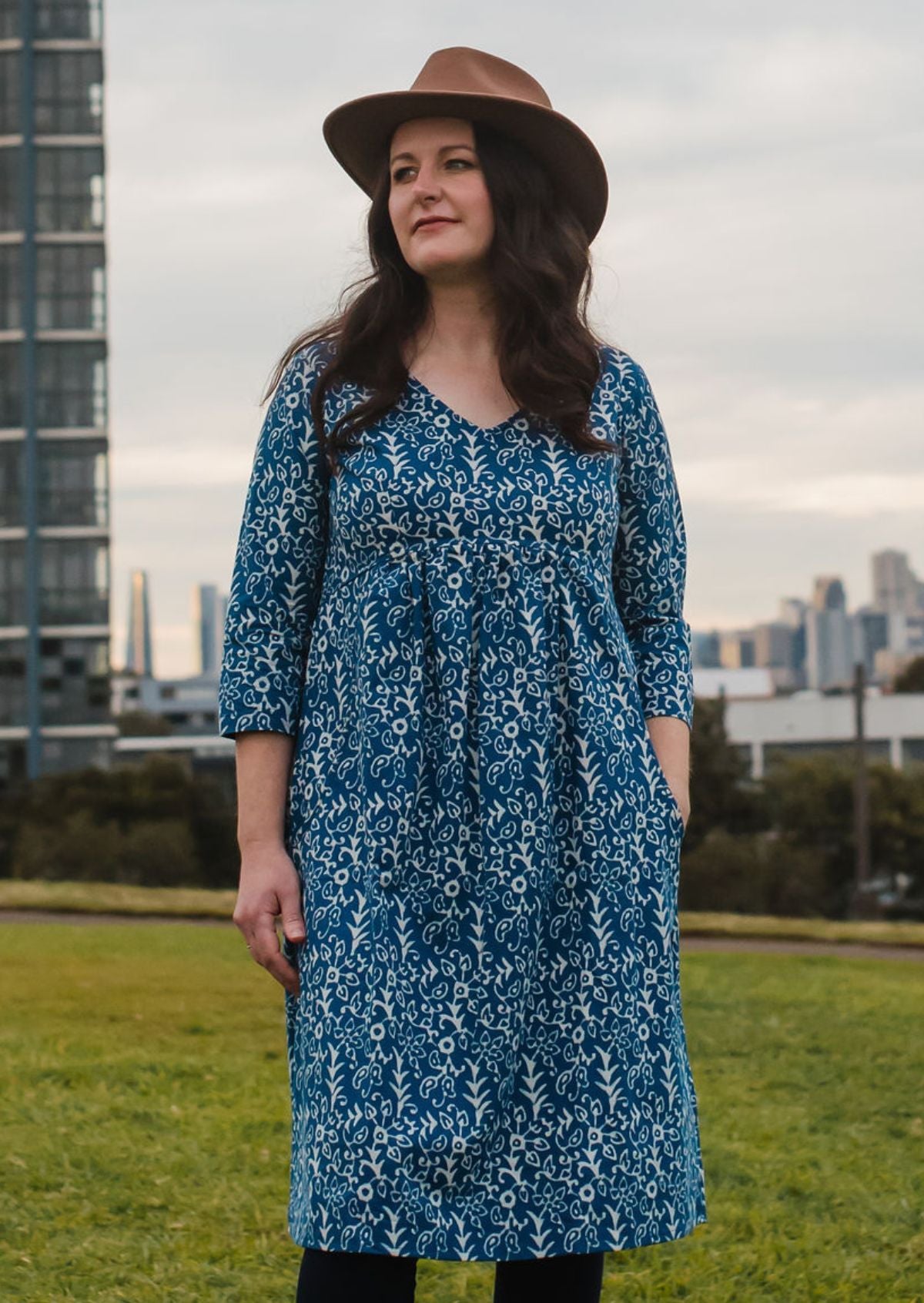 Woman wearing a blue floral dress and brown hat standing in an outdoor setting with buildings in the background.