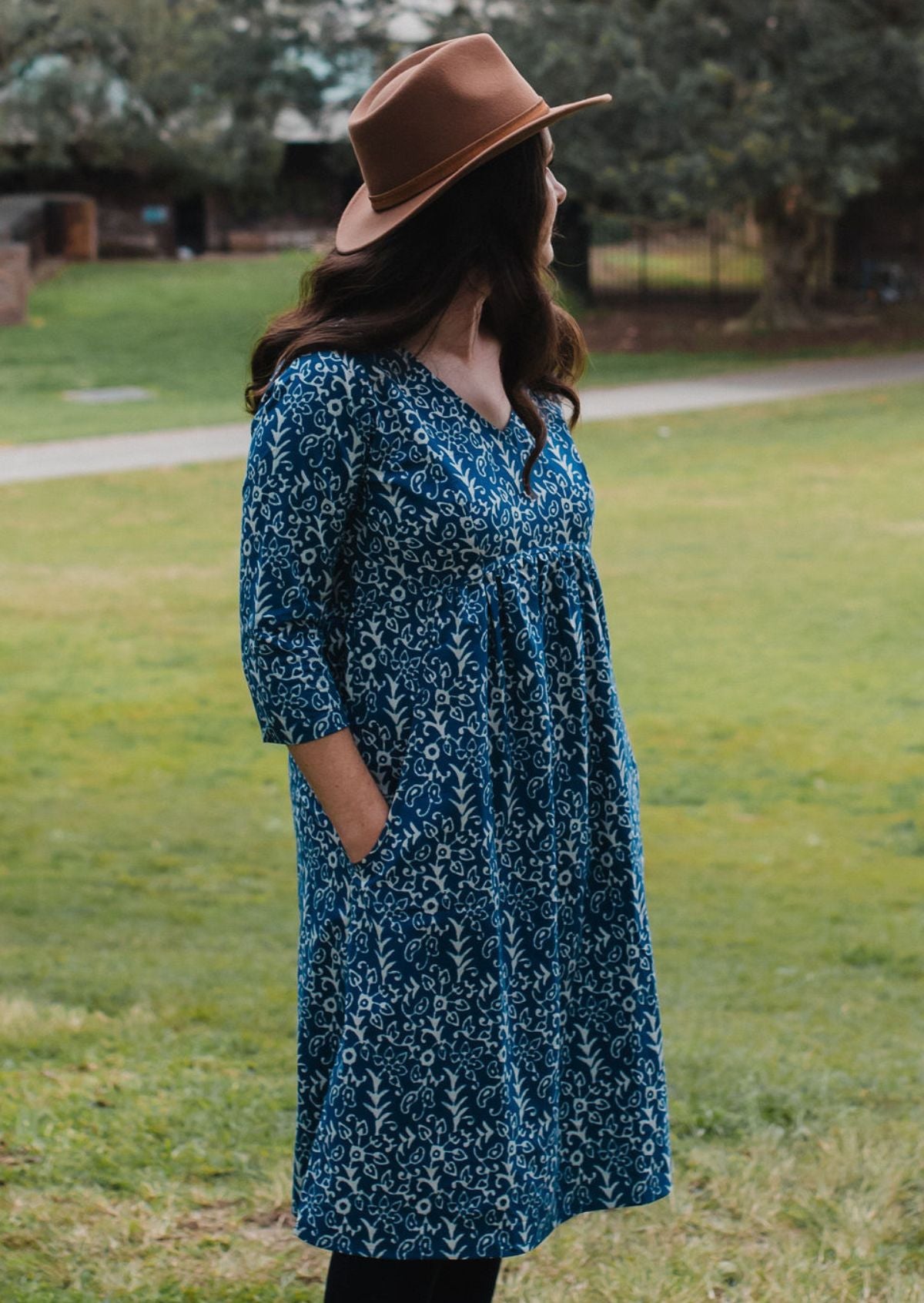 Woman wearing a blue floral dress and brown hat standing in a grassy area.