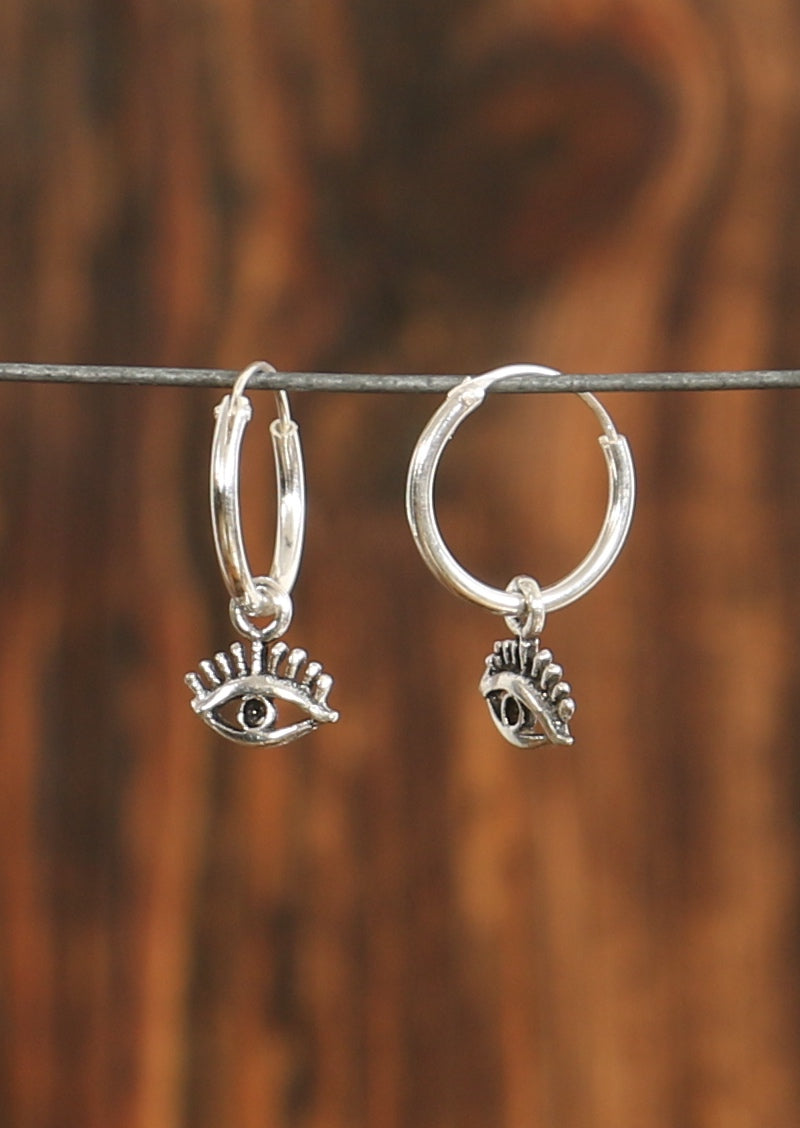 A pair of silver hoop Mystical Eye earrings with dangling eye-shaped charms, displayed against a wooden background.