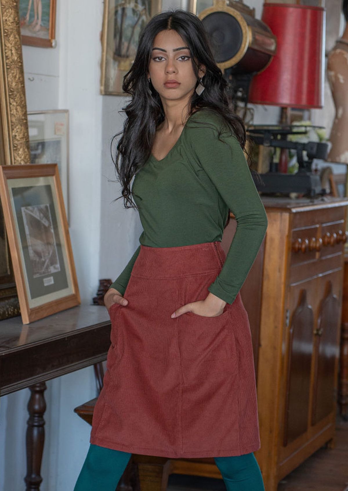 Model wears corduroy A-line knee length skirt with hands in pockets, paired with an olive top and teal leggings
