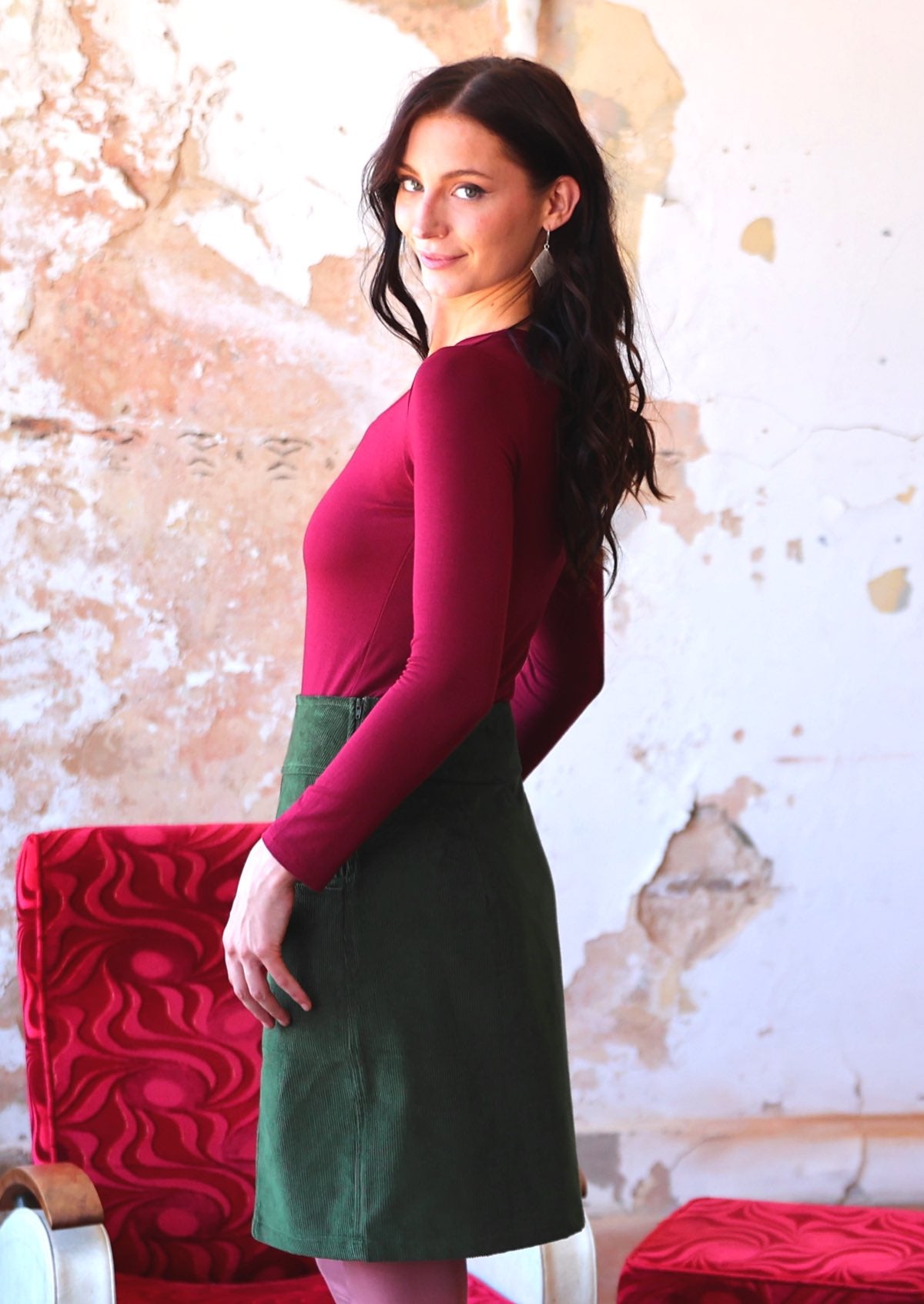 Woman stands side on in a green corduroy skirt with a side zip, paired with a bright maroon long sleeve top