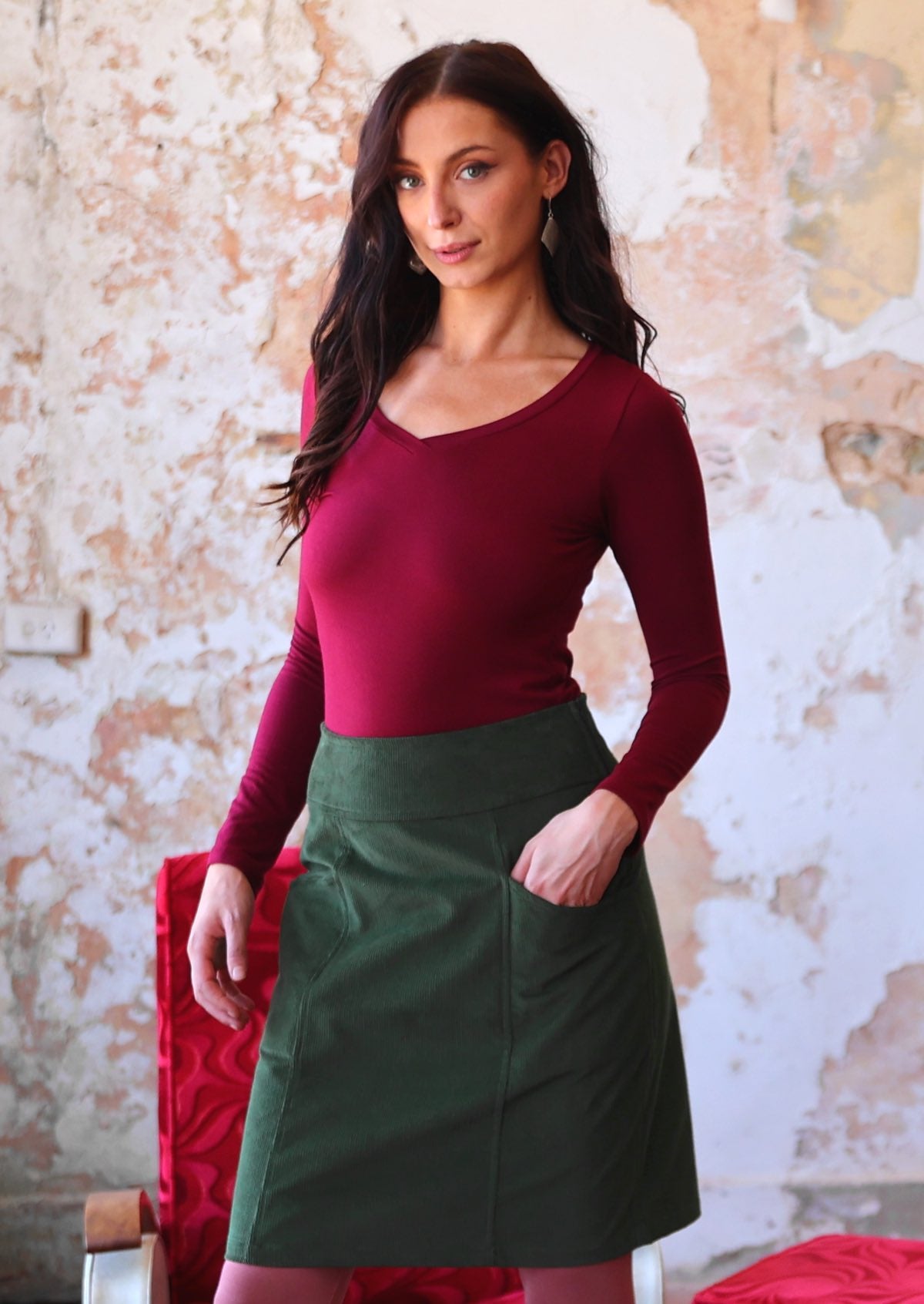 Woman poses in A-line green corduroy skirt with wide waistband, and with one hand in pocket