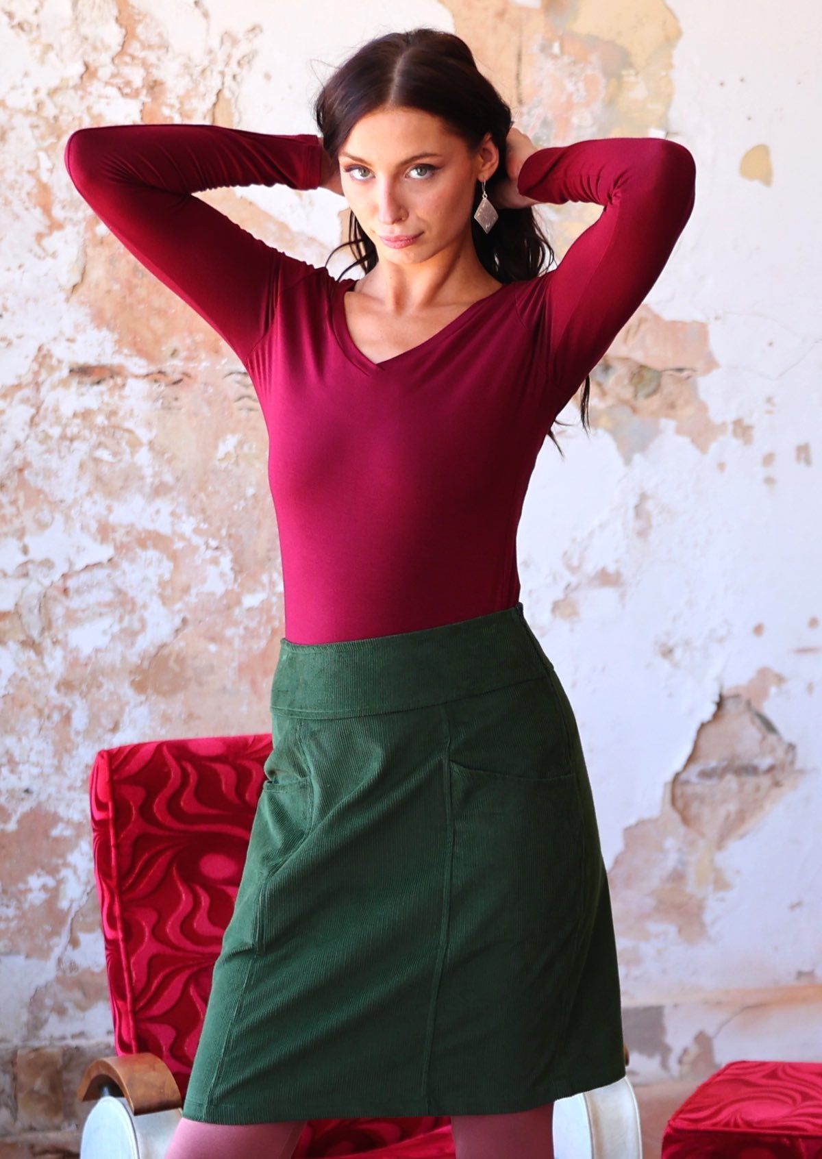Woman stands with hands above her head, in a green corduroy A-line above knee skirt paired with a maroon long sleeve top