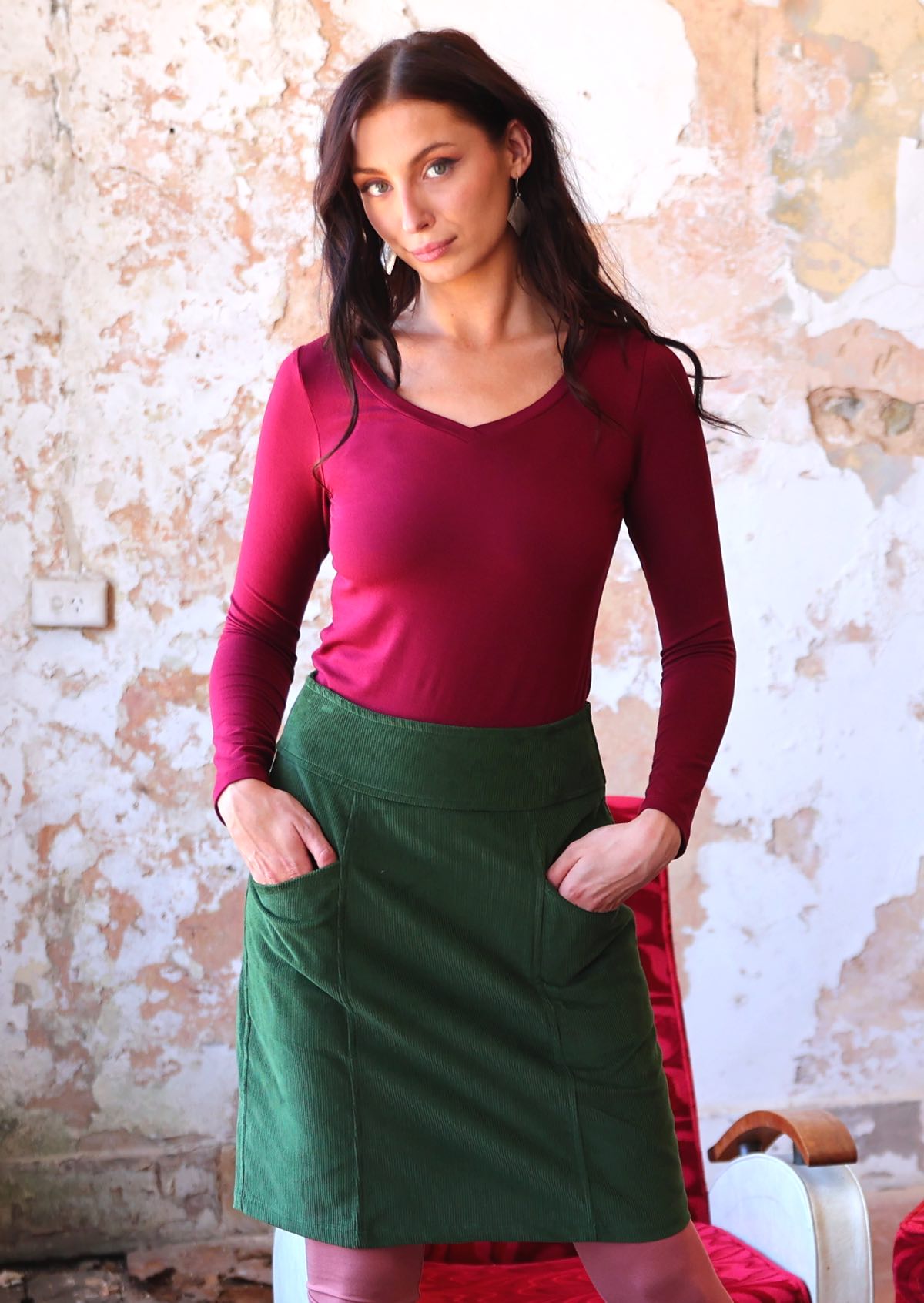 Woman wears green corduroy A-line skirt with her hands in pockets, paired with a maroon long sleeve V-neck top