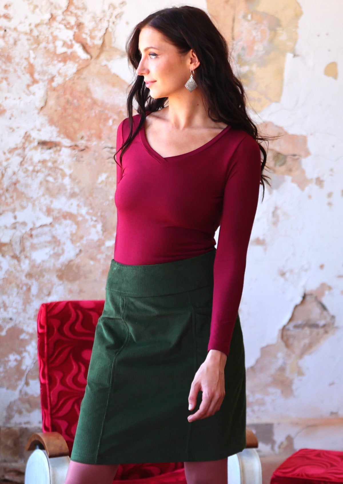 Woman wears a green corduroy A-line skirt with wide waistband and side zip, paired with a maroon long sleeve top