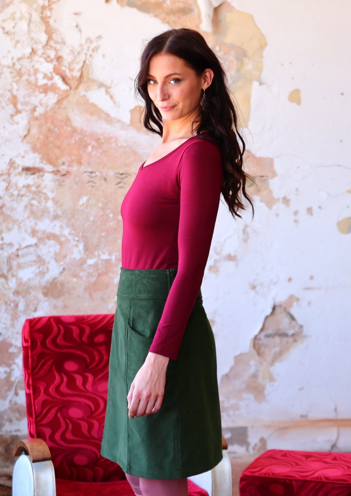 Model stands side on in green corduroy above knee skirt, paired with maroon long sleeve top