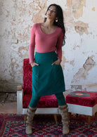Woman wears cotton corduroy A-line knee length skirt in teal with hands in pockets, paired with a pink long sleeve top, leggings and boots