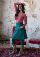 Woman poses in teal corduroy A-line knee length skirt with hands in pocket, paired with a pink top