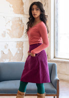 Model poses in plum purple corduroy knee length skirt with side zip and her hands in pockets