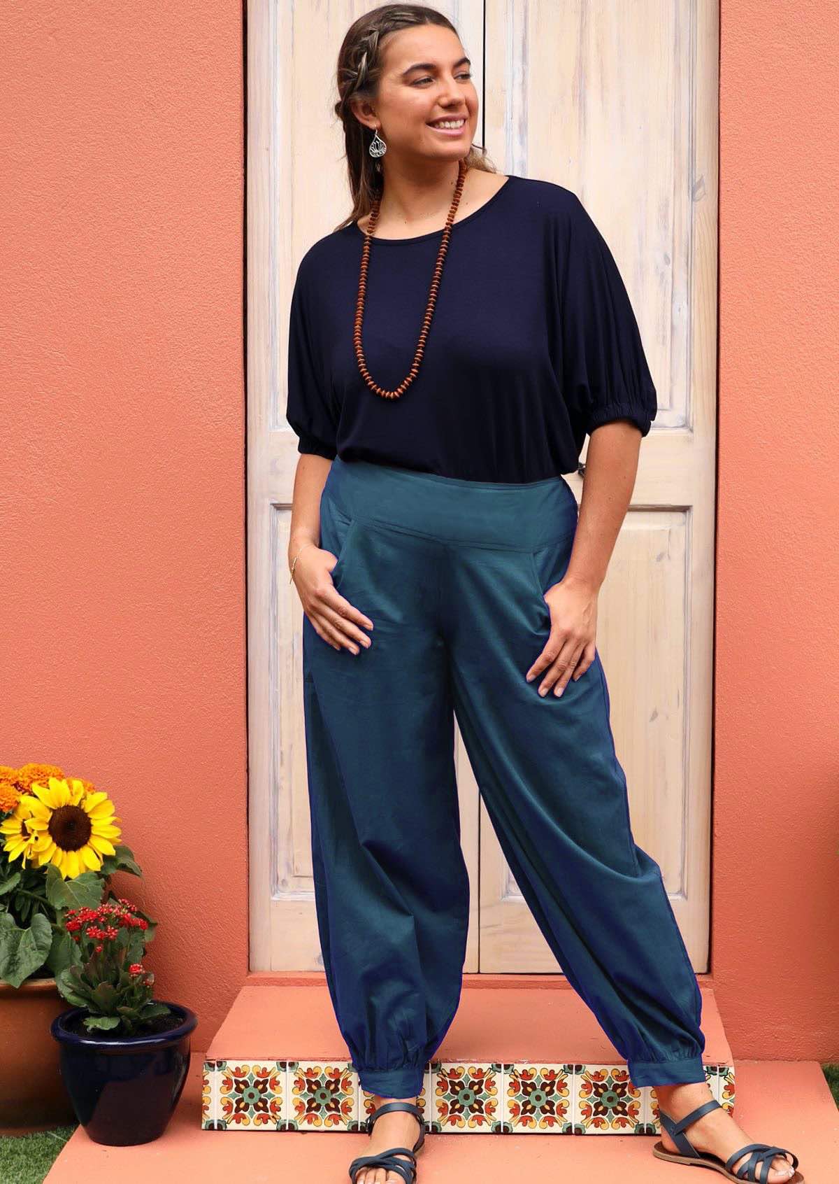 Woman stands in blue cotton harem pants with cuffed ankles and wide waistband, with her thumbs in pockets