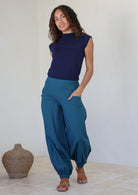 Woman stands with one hand in pocket wearing a genie style pull on pants in deep teal with cuffed ankles, wide leg fit and flat wide waist band that is shirred at the back