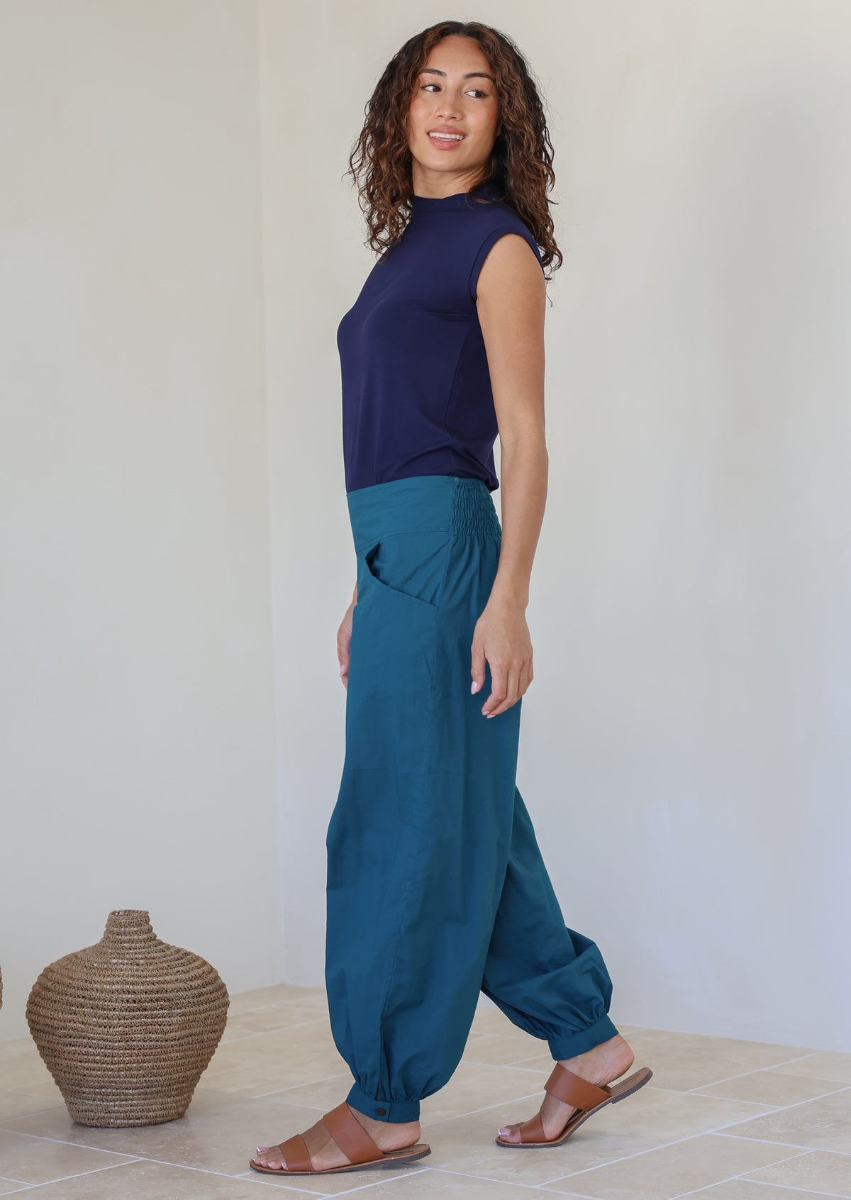 Woman wearing a navy top and relaxed fit teal cotton pants with cuffed, buttoned ankles and pockets, standing against a plain wall with a basket on the floor.