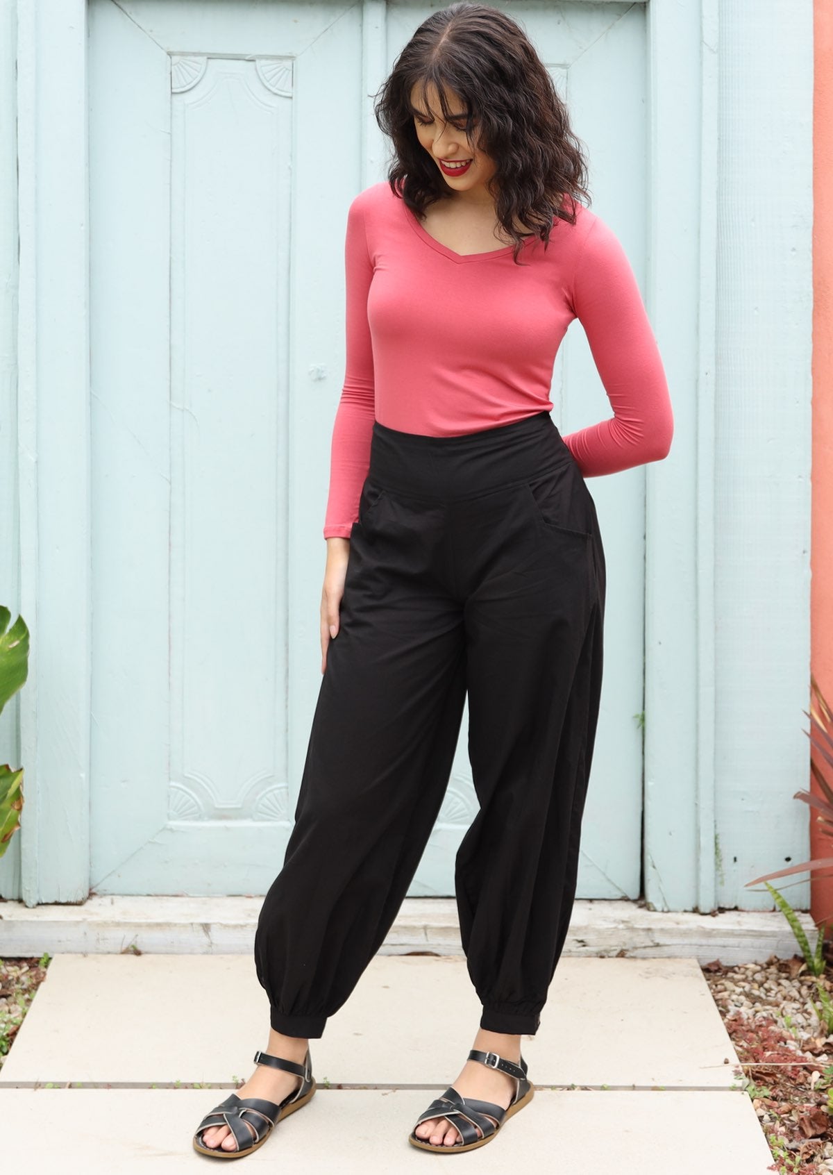 Woman wears black cotton pants with wide waistband and buttoned ankle cuff in fornt of pale blue doors