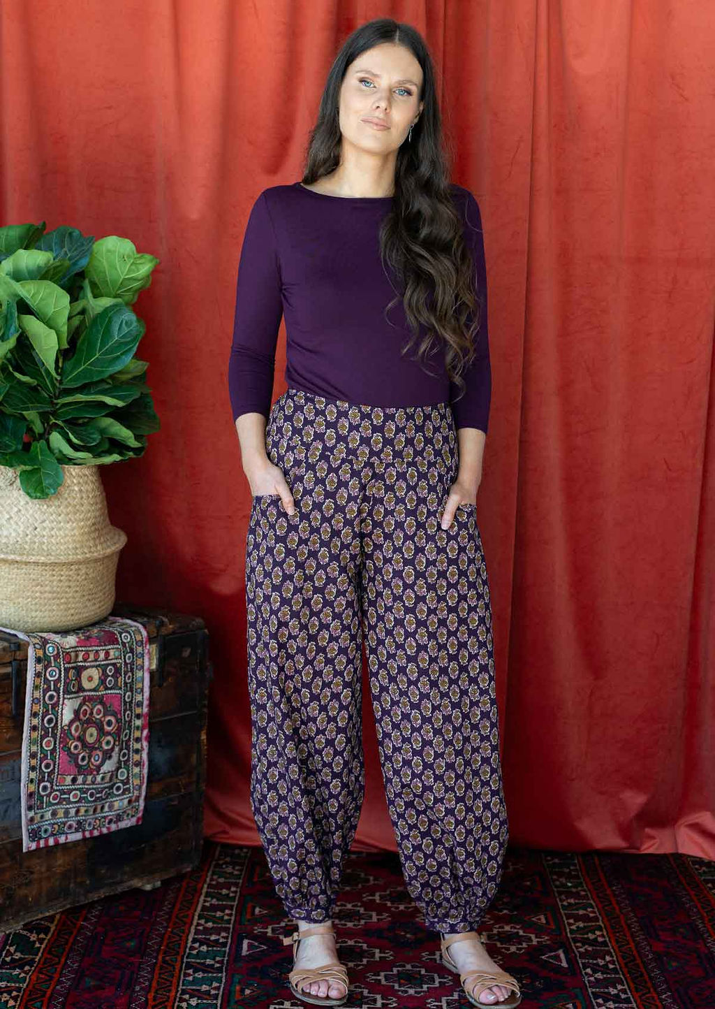 Woman wears cotton floral print with dark purple base harem pants, with buttoned ankle and pockets, paired with a dark purple top