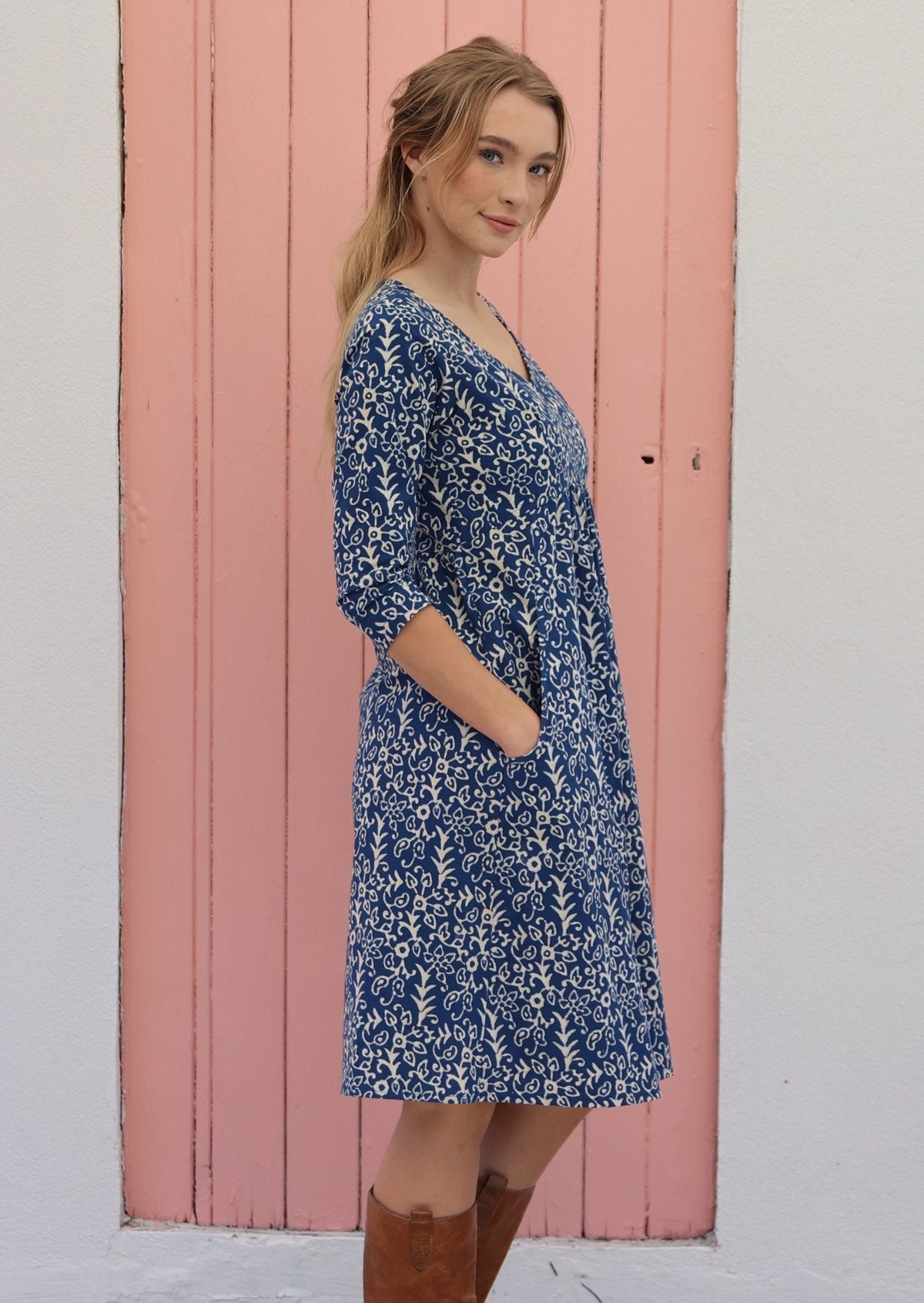Woman wears cotton over the knee length dress with 3/4 sleeves and V-neckline, with hands in pockets