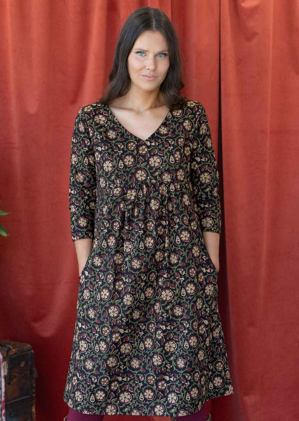 Woman wears cotton relaxed fit dress with a V-neckline and gathers in the empire waistline, with her hands in the deep pockets