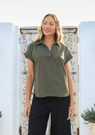Woman wearing an olive green shirt style relaxed fit top and black pants standing in front of a decorative white wall.