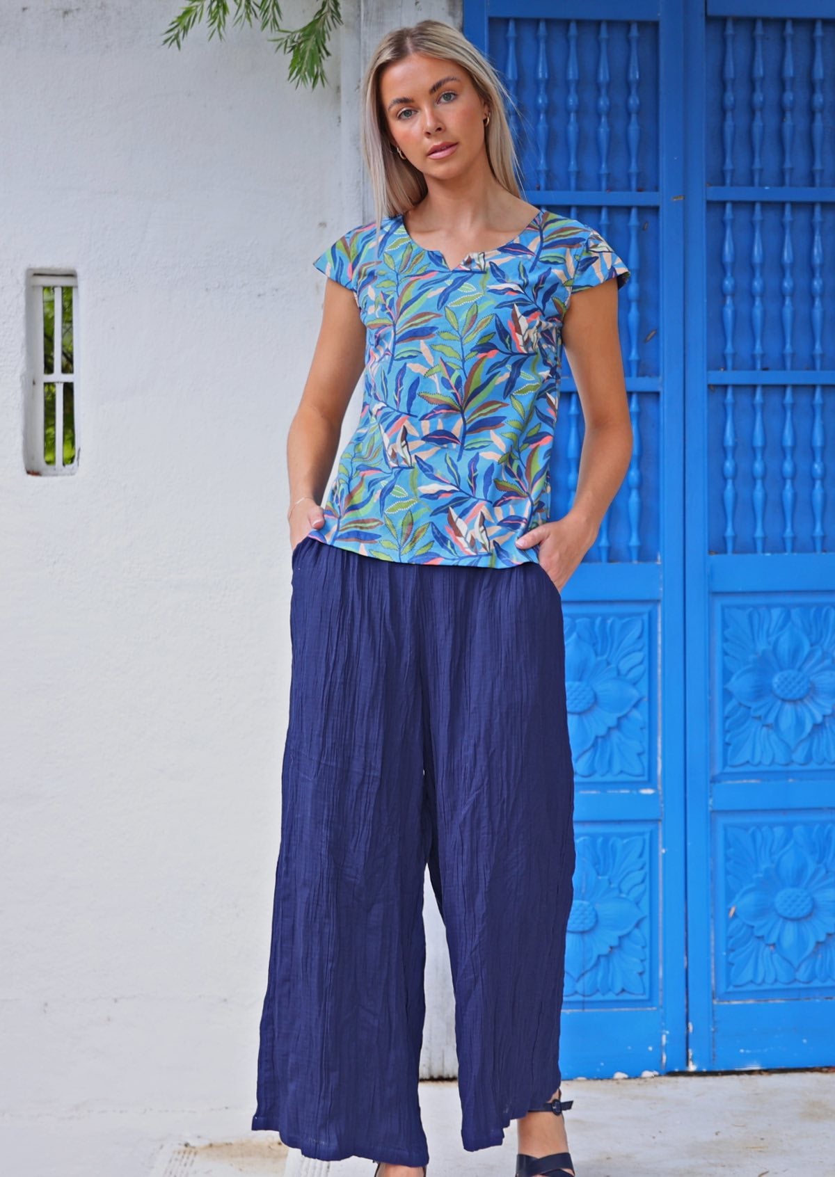 Woman standing in front of blue door wearing cap sleeved cotton tailored top in multicoloured leaf print on a blue base with hands in pants pockets