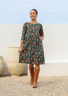 Woman wears 100% cotton knee length dress in deep green with beautiful pinkish red floral print and hand in pocket.