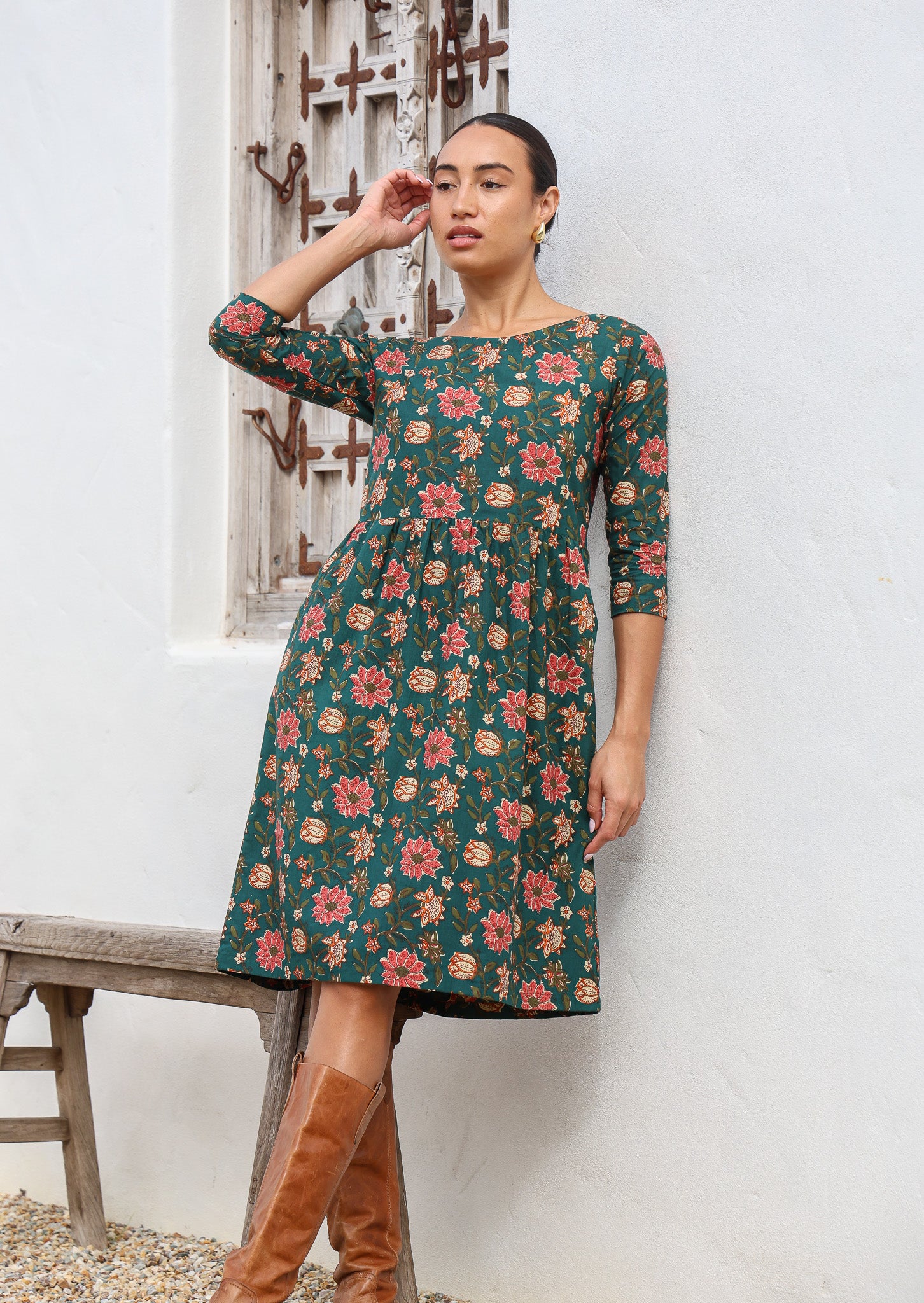 Woman in midlength cotton dress in dark green based floral print with 3/4 sleeves, wide round neckline and pockets.