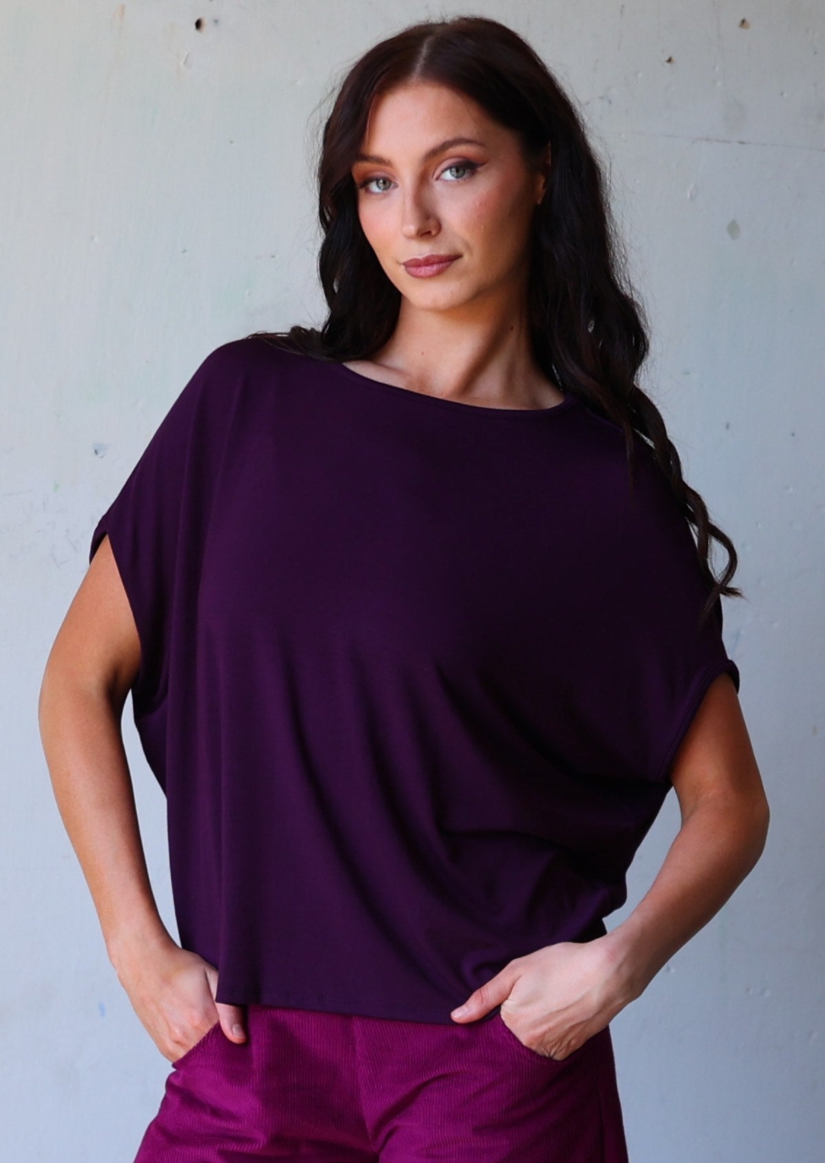 woman with dark hair wearing purple rayon batwing t-shirt and purple corduroy pants with hands in pockets