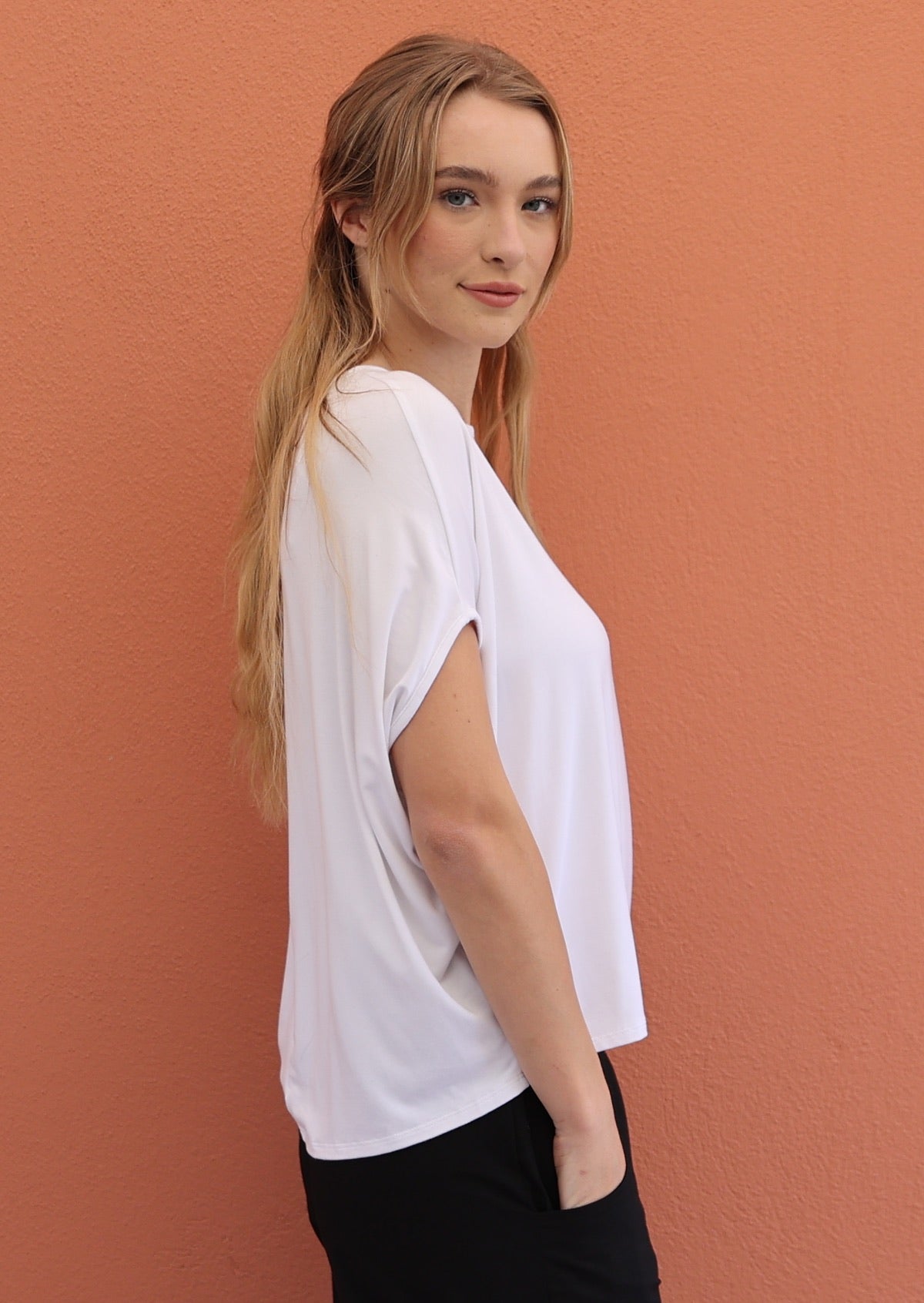 Woman stands side-on in white short sleeve batwing rayon top with hand in pocket of black pants