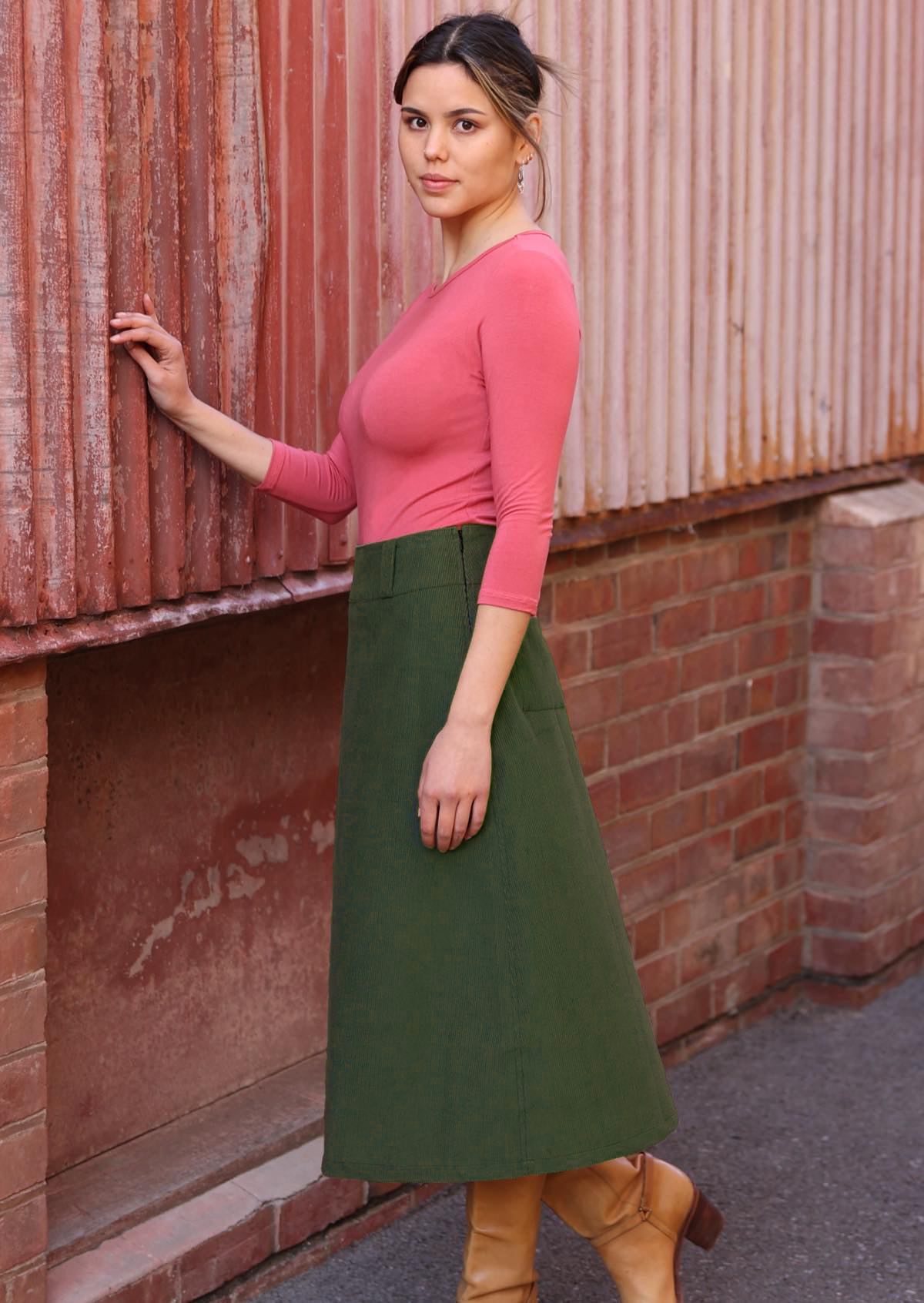 Woman stands side on in A-line midi length corduroy skirt in green with belt loops and a side zip, paired with a pink top