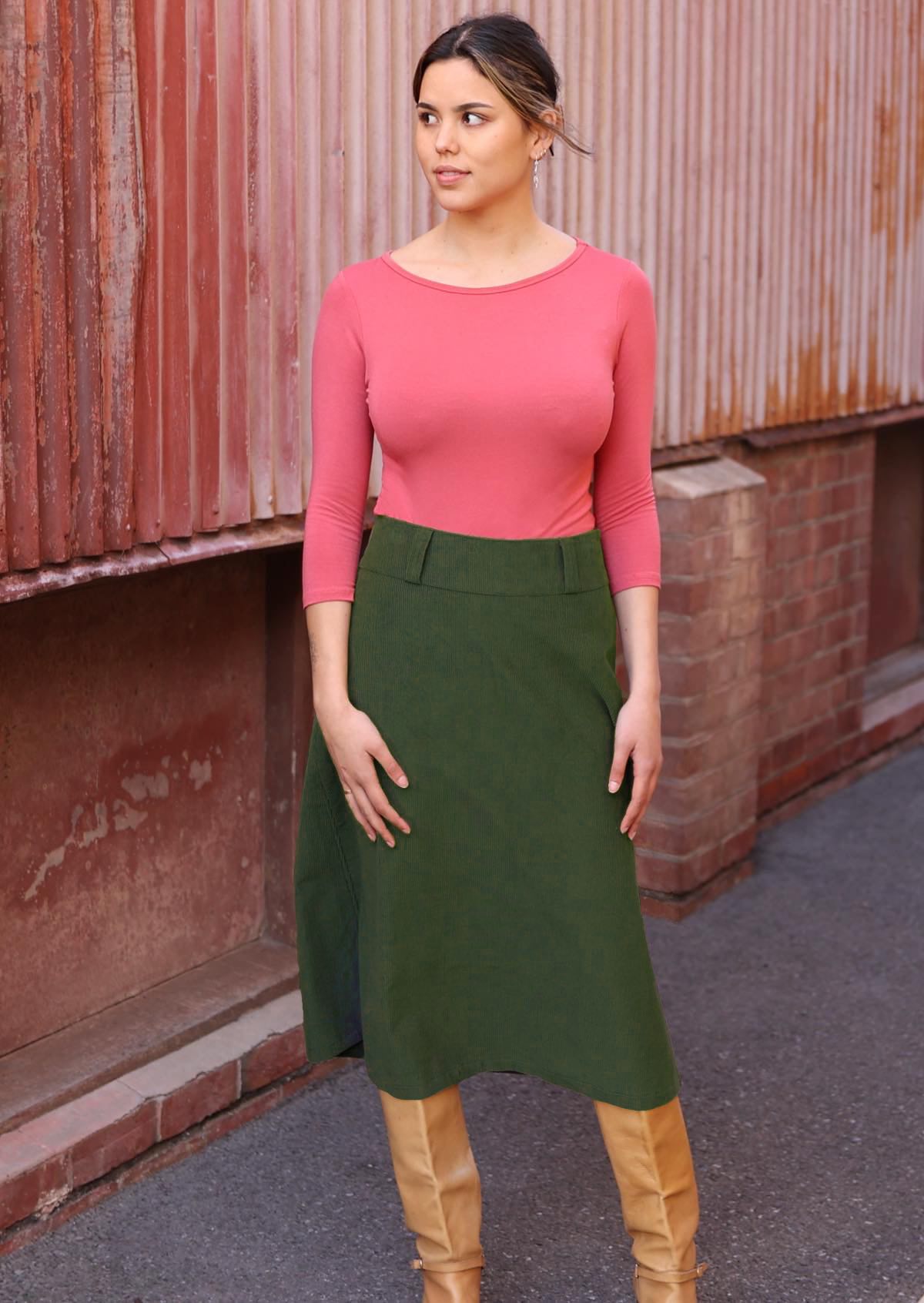Woman wears a midi length green corduroy skirt with belt loops, paired with a pink top