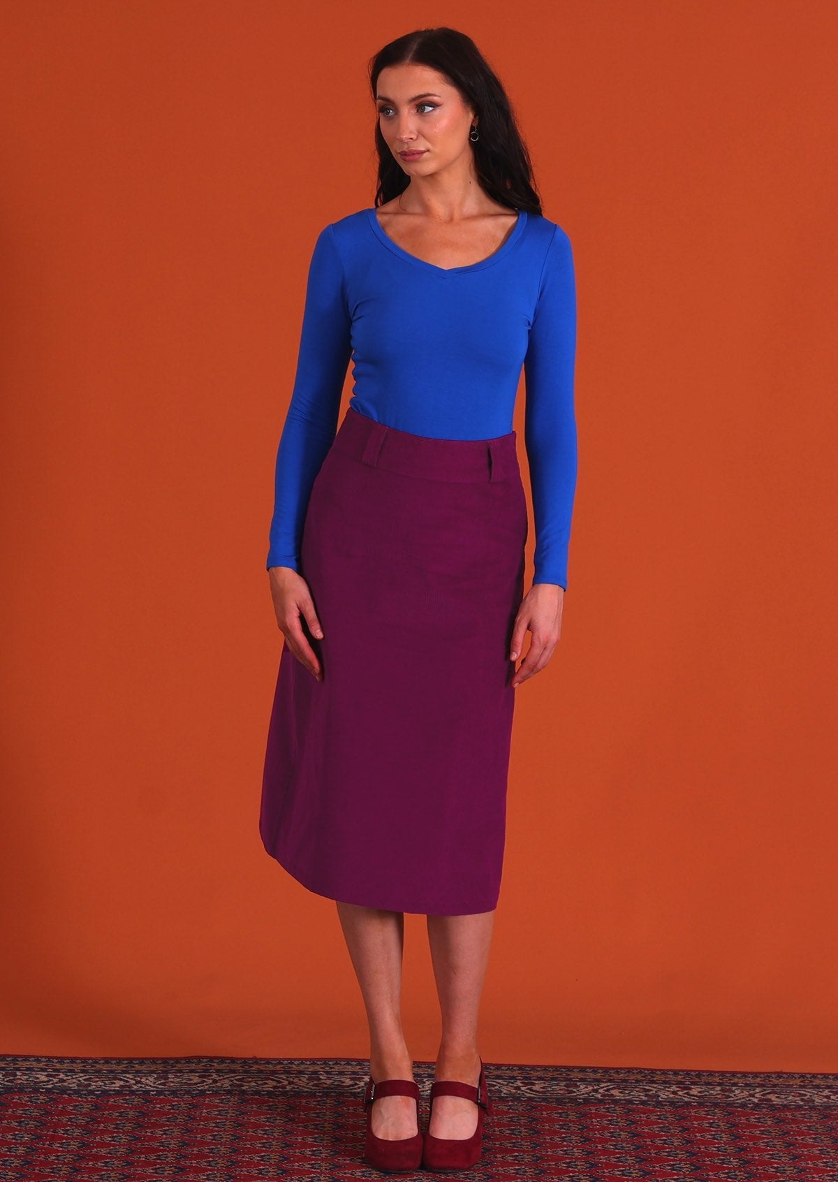 woman wearing purple corduroy aline skirt  with belt loops with bright blue long sleeve top standing on persian carpet