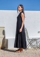 Woman in a long black sundress standing outdoors against a white wall with blue sky.