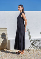 Woman in a black maxi dress standing outdoors against a white wall with a clear blue sky.