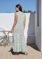 Woman wearing a white cotton patterned dress standing outdoors with a white wall and chair in the background.