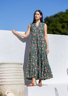 Woman wearing a green floral cotton maxi dress against a white wall with blue sky and greenery.