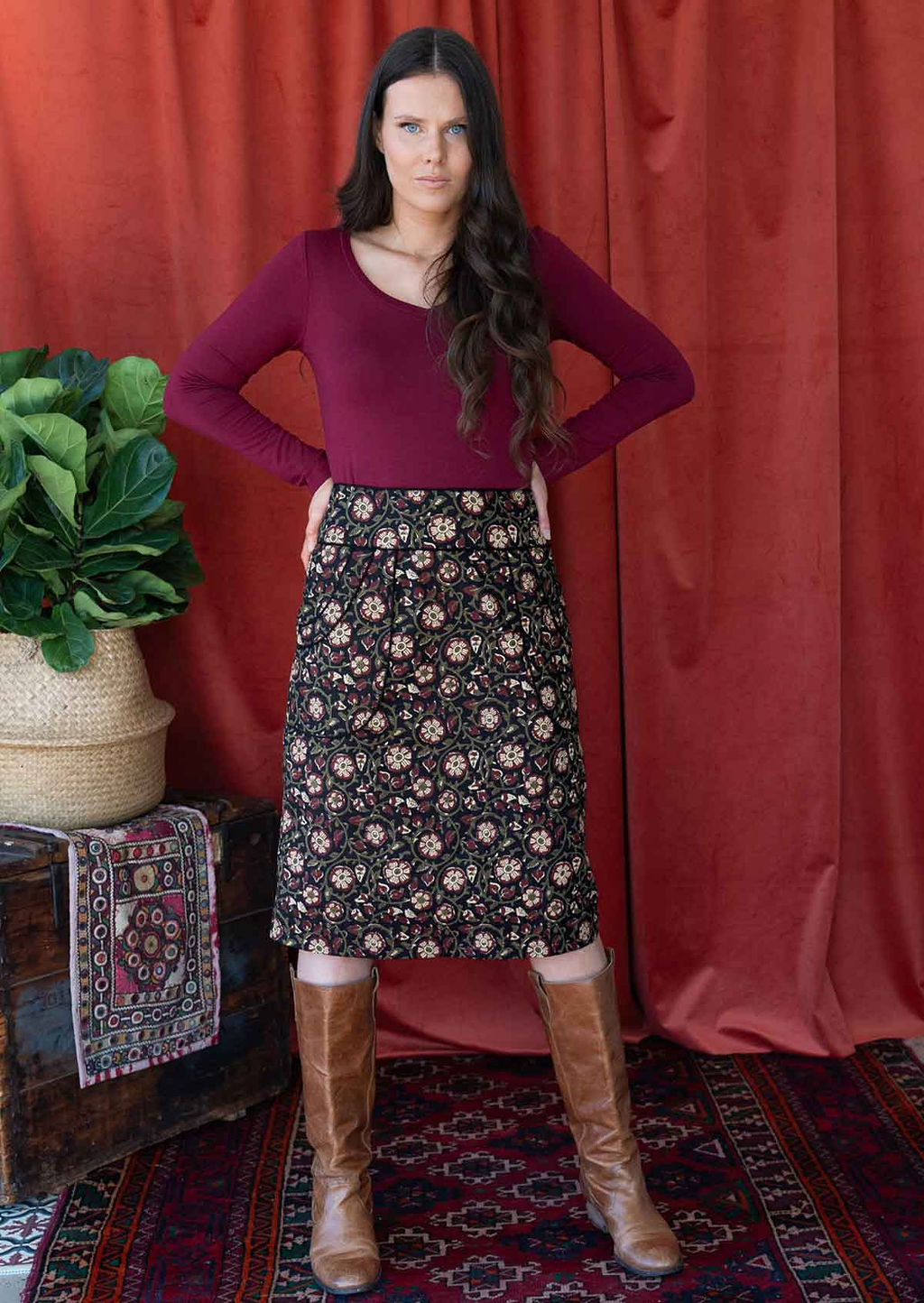 Woman stands in over the knee cotton pencil skirt with a floral print on a black base, paired with a maroon long sleeve top