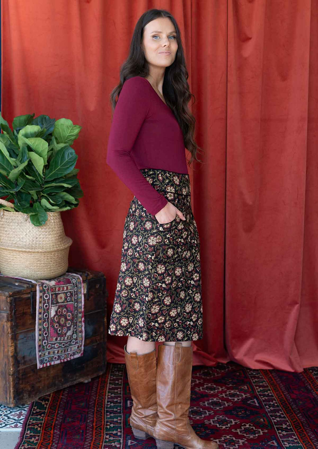 Model stands side on in black based cotton pencil skirt with high waistband paired with a maroon long sleeve top, with hands in pockets
