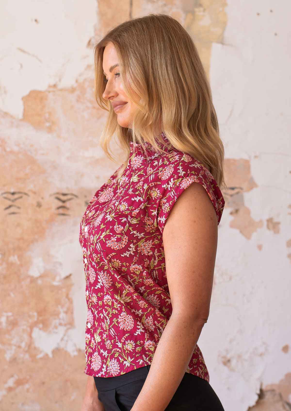 Woman stands sideways in cotton cap sleeve button through shirt with folded cuffs, in floral with bright red base