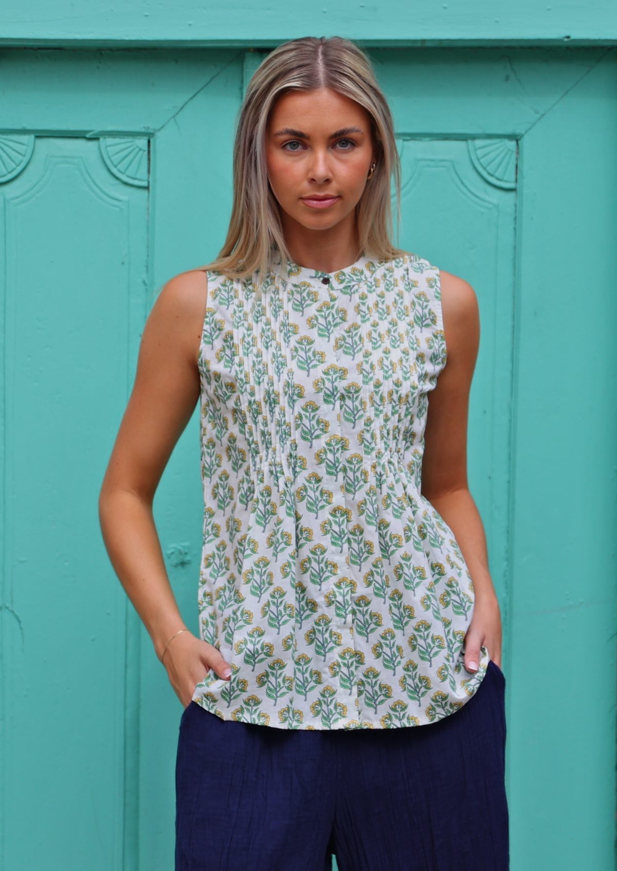 Model wears white based yellow and green floral print cotton sleeveless blouse with tiny decorative pleats and a high round neckline