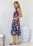 Woman wearing a floral midi length dress standing next to a textured vase against a white wall, with hands in pockets