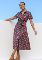Model wears shirt style cotton dress in bright floral print with fabric waist tie, pockets and buttons throughout the dress.