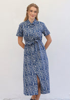 Woman wears cotton button through shirt dress with collar and short sleeves, in off white floral print on a blue base