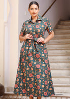 Woman poses in cotton green based floral print shirt style dress with thick fabric belt, buttons throughout the dress and pockets.