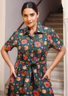 Model with hands in pocket wears green floral printed, button through dress with collar and removable waist tie.