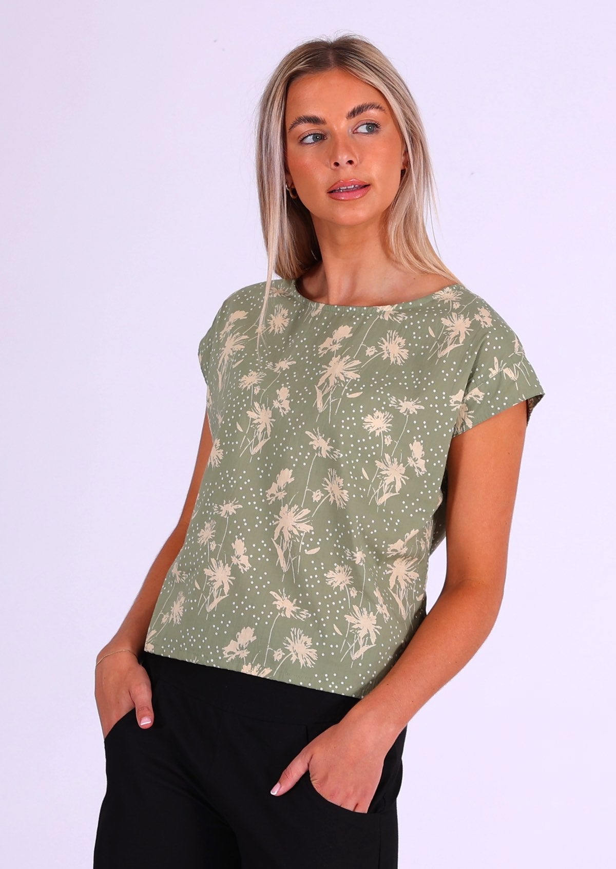 Model wears cotton short sleeve top with cream floral silhouettes on a pale green base, with hands in pockets of black cotton pants