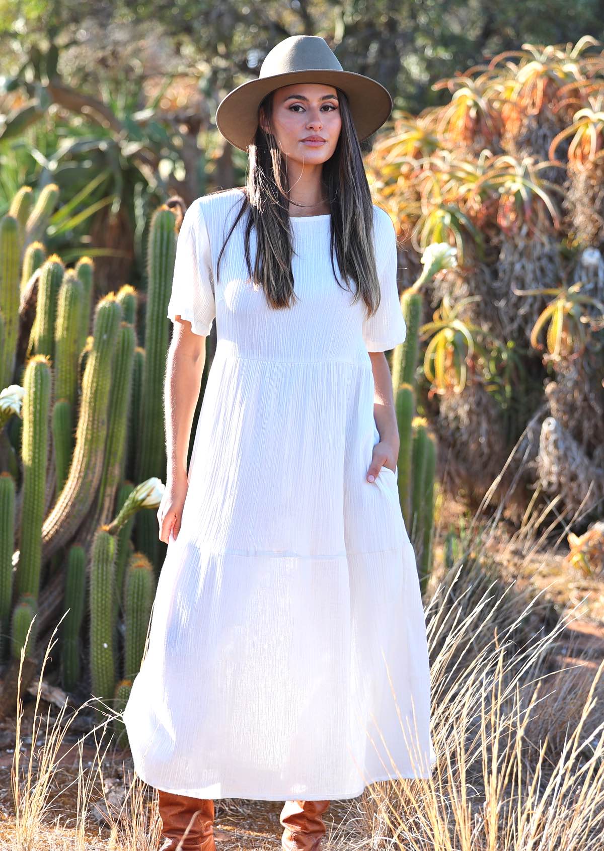 Woman with dark hair in beautiful
white cotton maxi dress with hands in pockets and hat on head