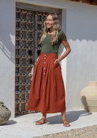 Woman standing with hands in pocket wearing a 100% cotton rust colored button through maxi skirt with box pleats and belt loops