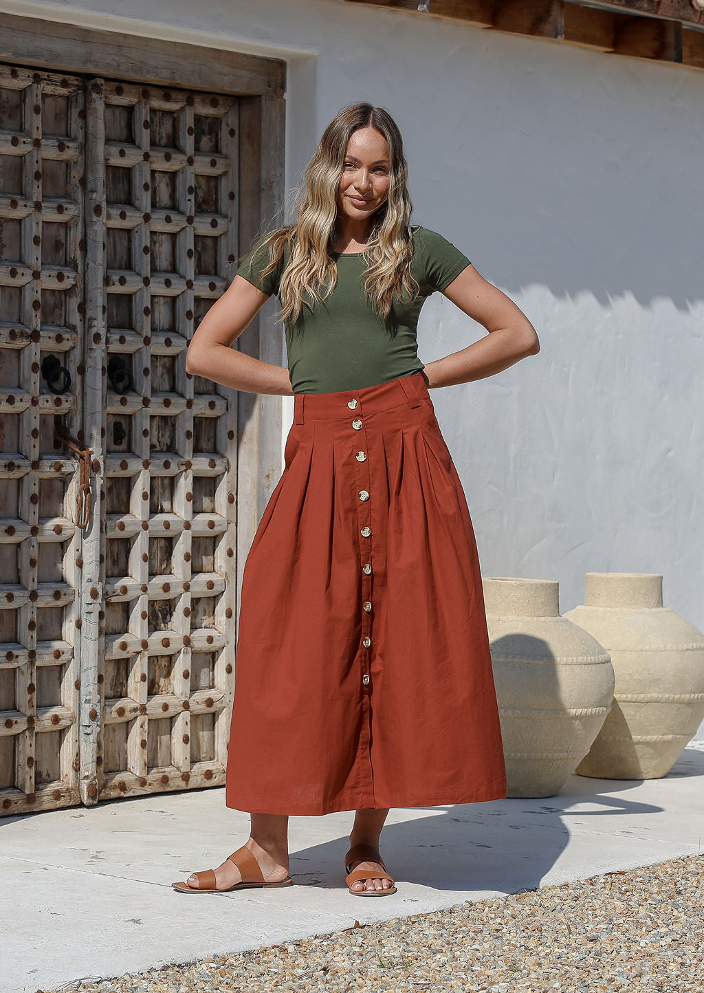 Woman wearing a green basic top and rust colored shin length, A line cotton skirt with box pleats, belt loops and button through front