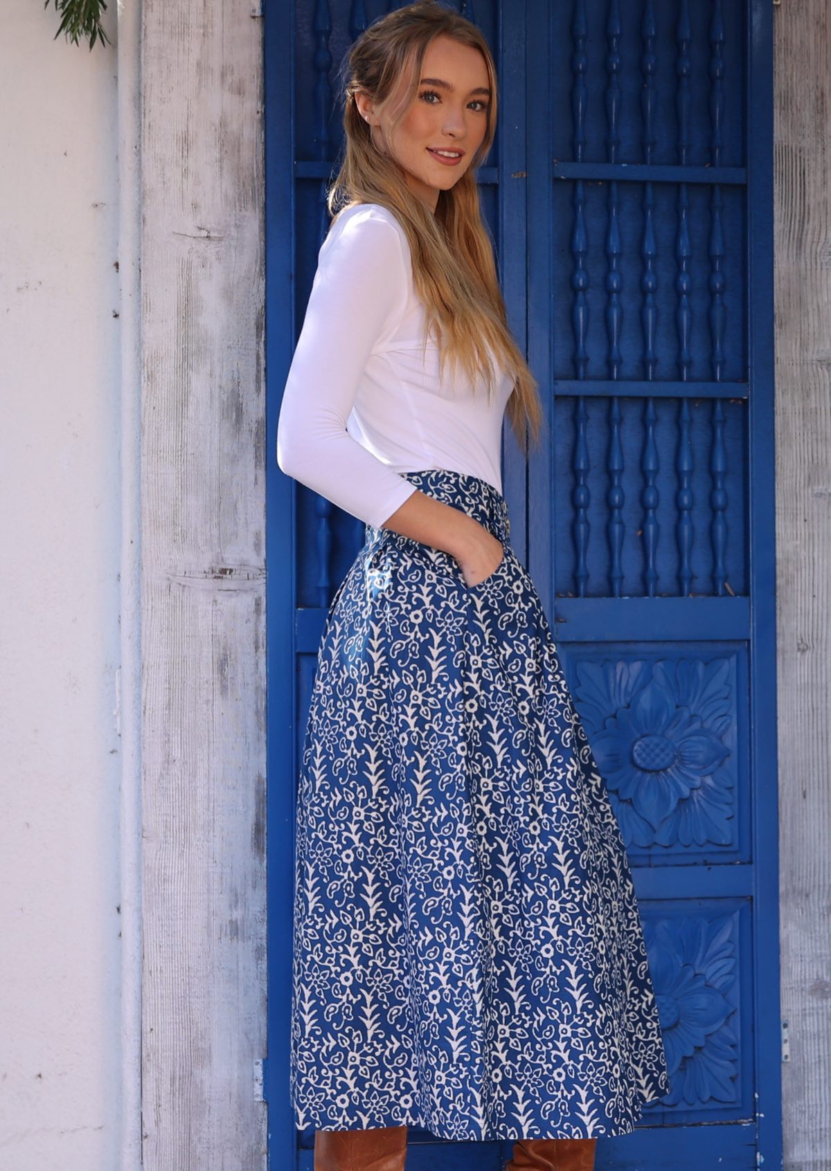 Woman stands sideways in midi length blue cotton skirt with white floral print, paired with white stretch rayon top