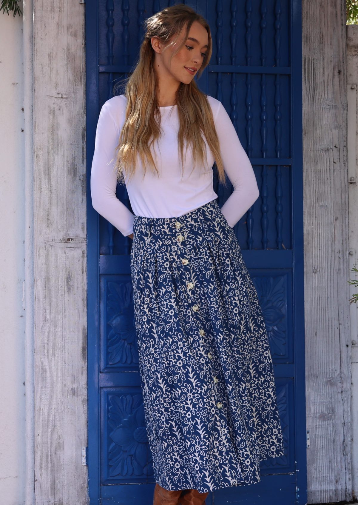 Woman leans against doors in blue cotton midi length button through skirt paired with a white stretch rayon top