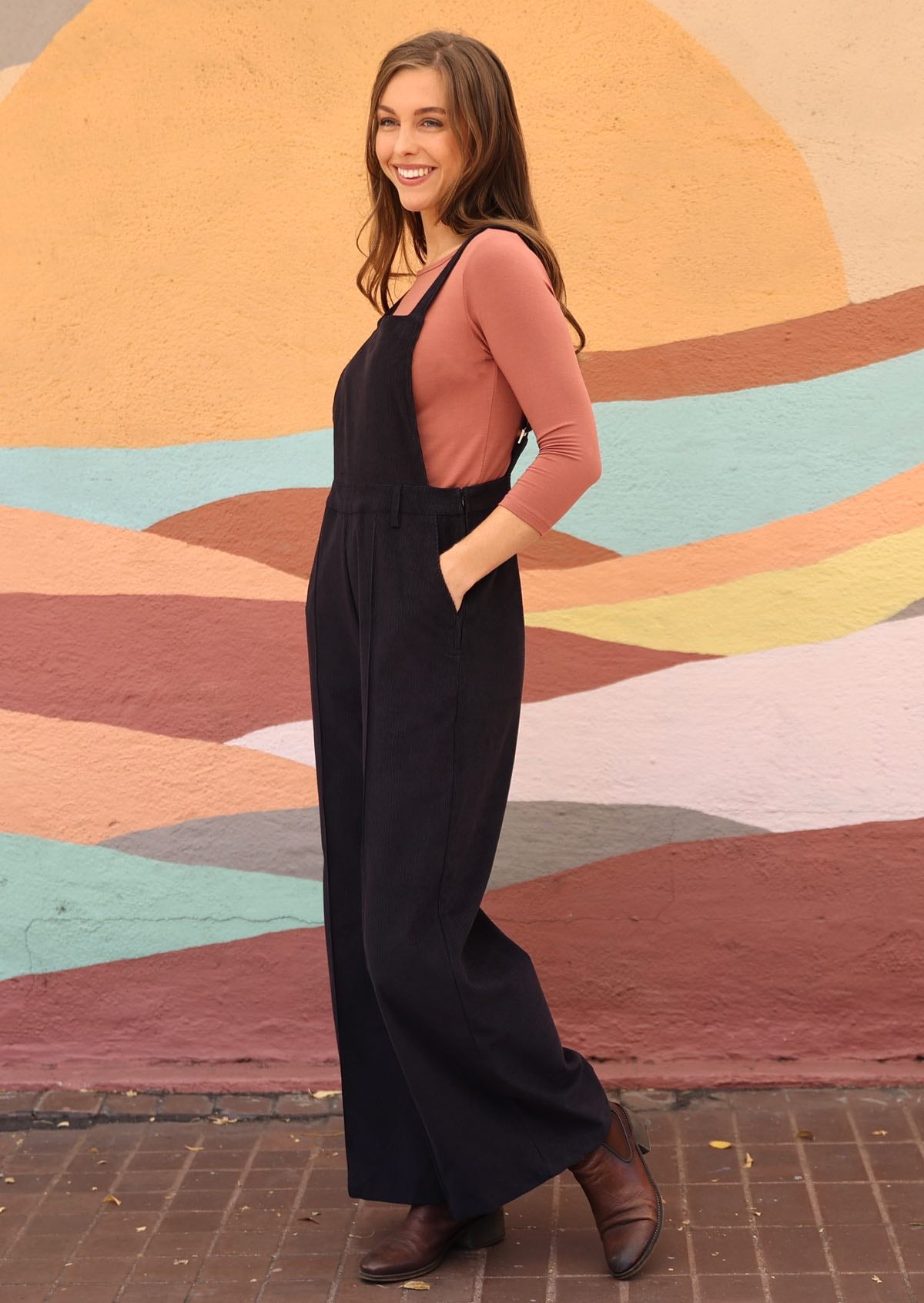 woman wearing cotton corduroy overalls in dark grey over dusty pink long sleeve top with hands in pockets
