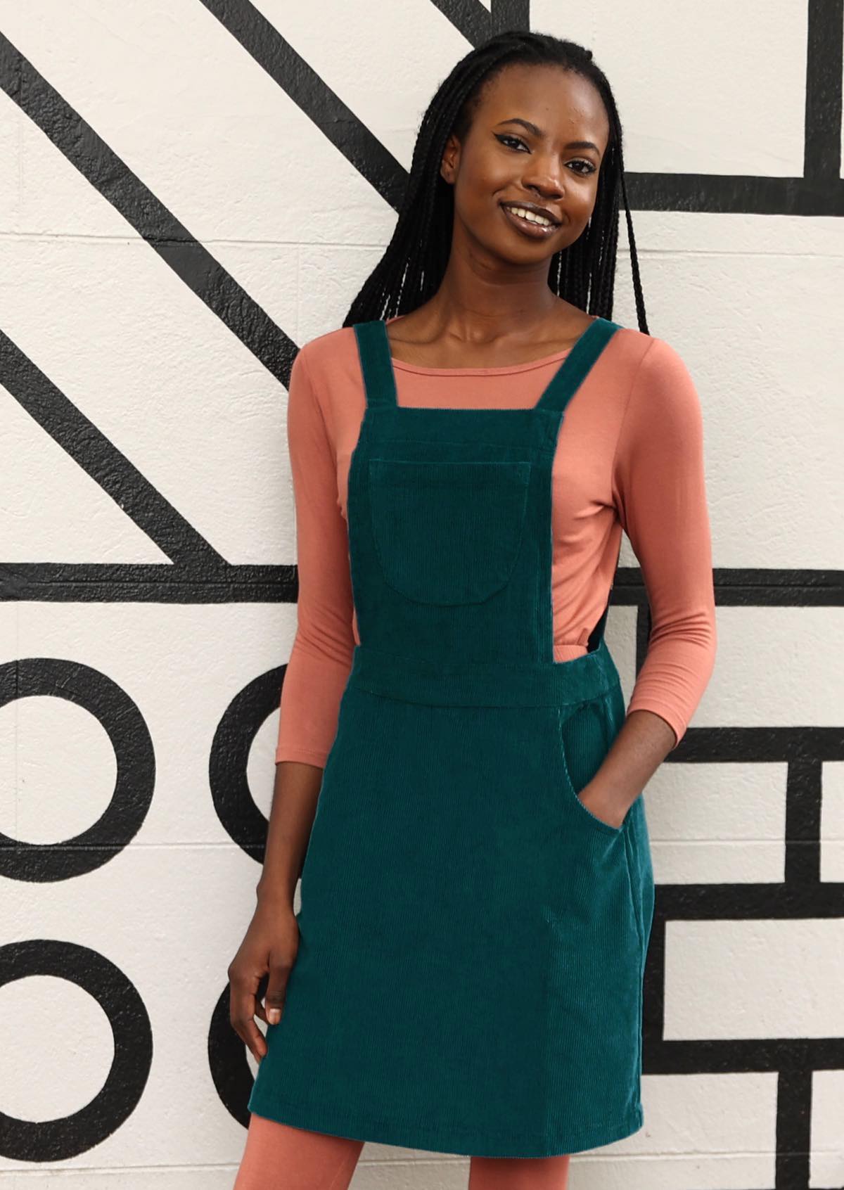 woman wearing teal corduroy pinafore over pink top, pink leggings and tan boots with hand in pocket